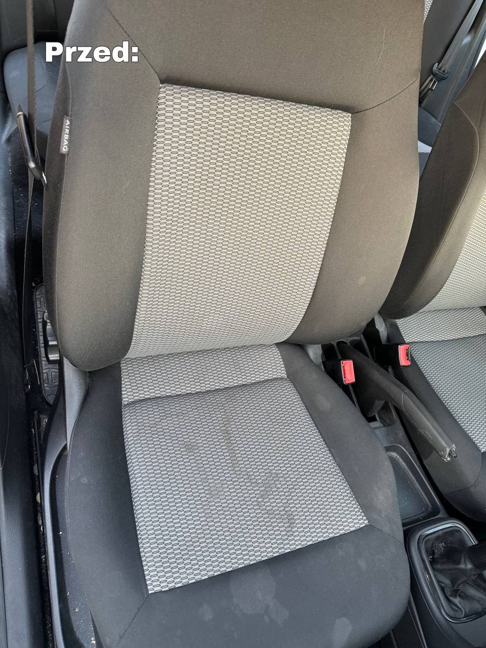 Brudne, szare siedzenie samochodowe z plamami przed czyszczeniem, widoczny fragment pasów bezpieczeństwa i dźwigni hamulca ręcznego.