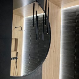 MACIEJ MATYSIAK - Nowoczesna łazienka z okrągłym lustrem, czarną armaturą i oświetleniem LED, ukazana pod kątem, eksponująca drewnopodobne i trójwymiarowe czarne płytki.