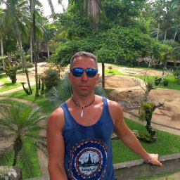 MACIEJ MATYSIAK - Mężczyzna w niebieskim podkoszulku z nadrukiem 'Bangkok' i odblaskowych okularach na tle tropikalnego ogrodu z palmami.