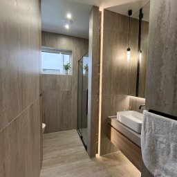 MACIEJ MATYSIAK - Nowoczesna łazienka w odcieniach beżu z kabiną prysznicową typu walk-in, prostokątną umywalką nablatową, podświetlanym lustrem i wiszącą lampą. Na parapecie okna doniczka z zieloną rośliną.