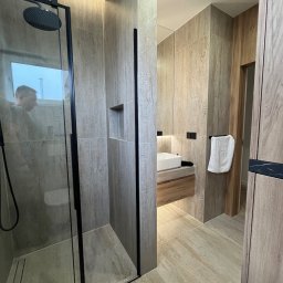 MACIEJ MATYSIAK - Nowoczesna łazienka z kabiną prysznicową typu walk-in z czarnymi ramami, umywalką nablatową na drewnianej szafce i beżowymi płytkami imitującymi trawertyn.