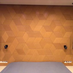 MACIEJ MATYSIAK - Ściana z korka 3D w geometryczne wzory, oświetlona wiszącymi czarnymi lampami i kinkietami, nad wezgłowiem łóżka z szarą pościelą.