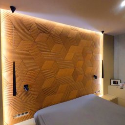 MACIEJ MATYSIAK - Nowoczesna sypialnia z drewnianą ścianą dekoracyjną z geometrycznym wzorem, podświetloną listwami LED, czarne lampki ścienne, łóżko z szarą pościelą, biała szafka nocna z dekoracjami.