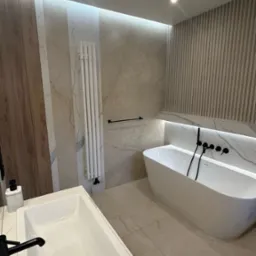 Nowoczesna łazienka z wolnostojącą wanną, białą umywalką nablatową z czarną armaturą i pionowym grzejnikiem drabinkowym na tle beżowych płytek imitujących marmur.