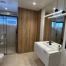 MACIEJ MATYSIAK - Nowoczesna łazienka z kabiną prysznicową typu walk-in z czarnymi ramami, drewnianą zabudową meblową, podwieszaną szafką z umywalką z blatem imitującym marmur i minimalistycznym oświetleniem.