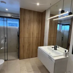 Nowoczesna łazienka z kabiną prysznicową typu walk-in z czarnymi ramami, drewnianą zabudową meblową, podwieszaną szafką z umywalką z blatem imitującym marmur i minimalistycznym oświetleniem.