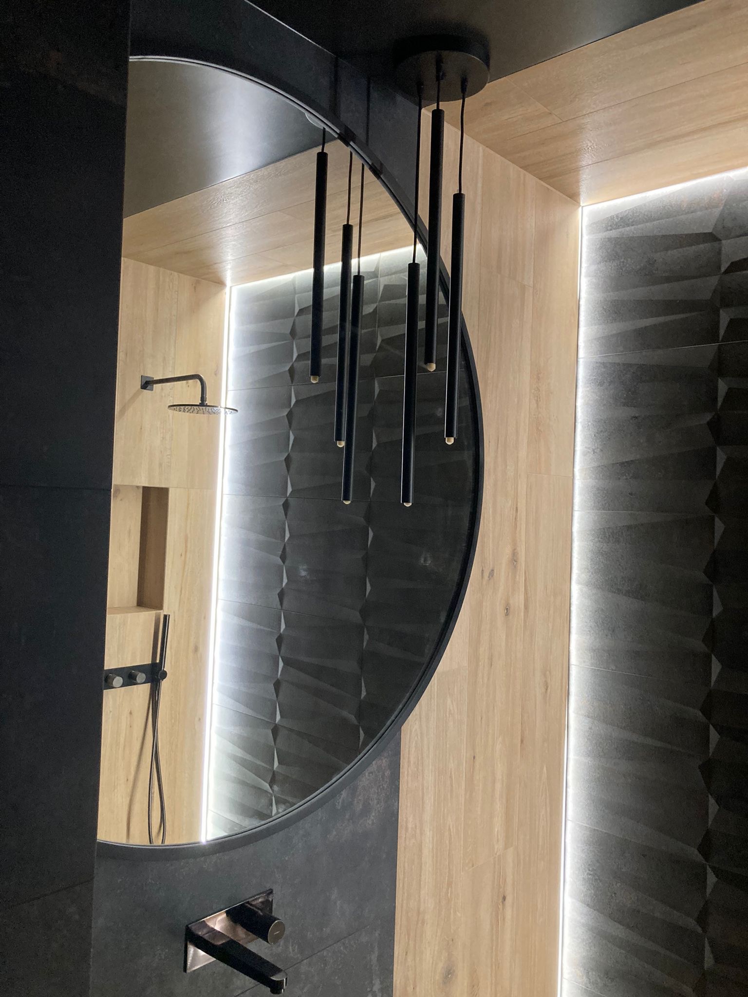 Nowoczesna łazienka z okrągłym lustrem, czarną armaturą i oświetleniem LED, ukazana pod kątem, eksponująca drewnopodobne i trójwymiarowe czarne płytki.
