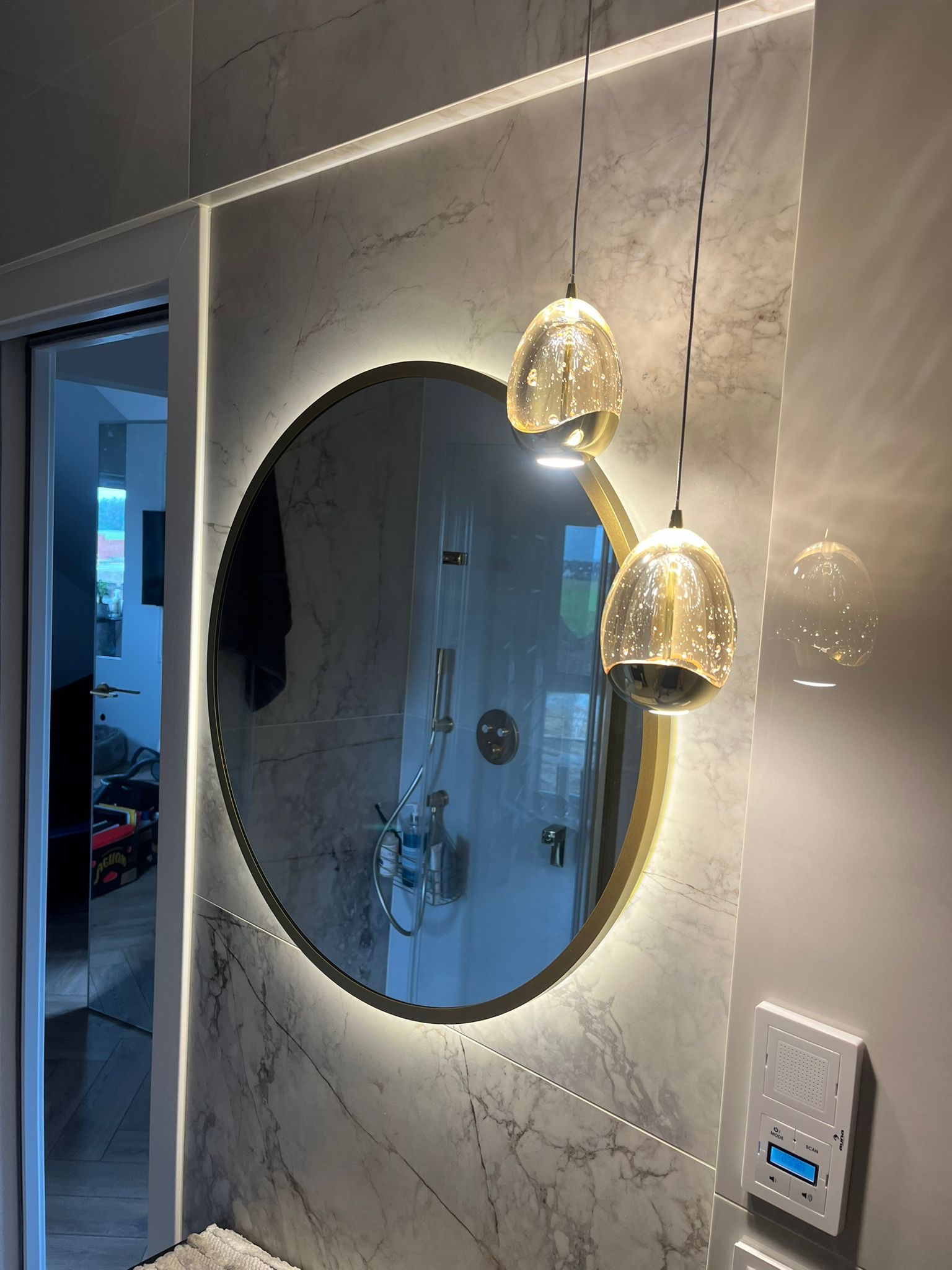 Okrągłe lustro z podświetleniem LED w łazience wykończonej płytkami imitującymi marmur, wiszące szklane lampy nad lustrem, widoczny panel sterowania na ścianie.
