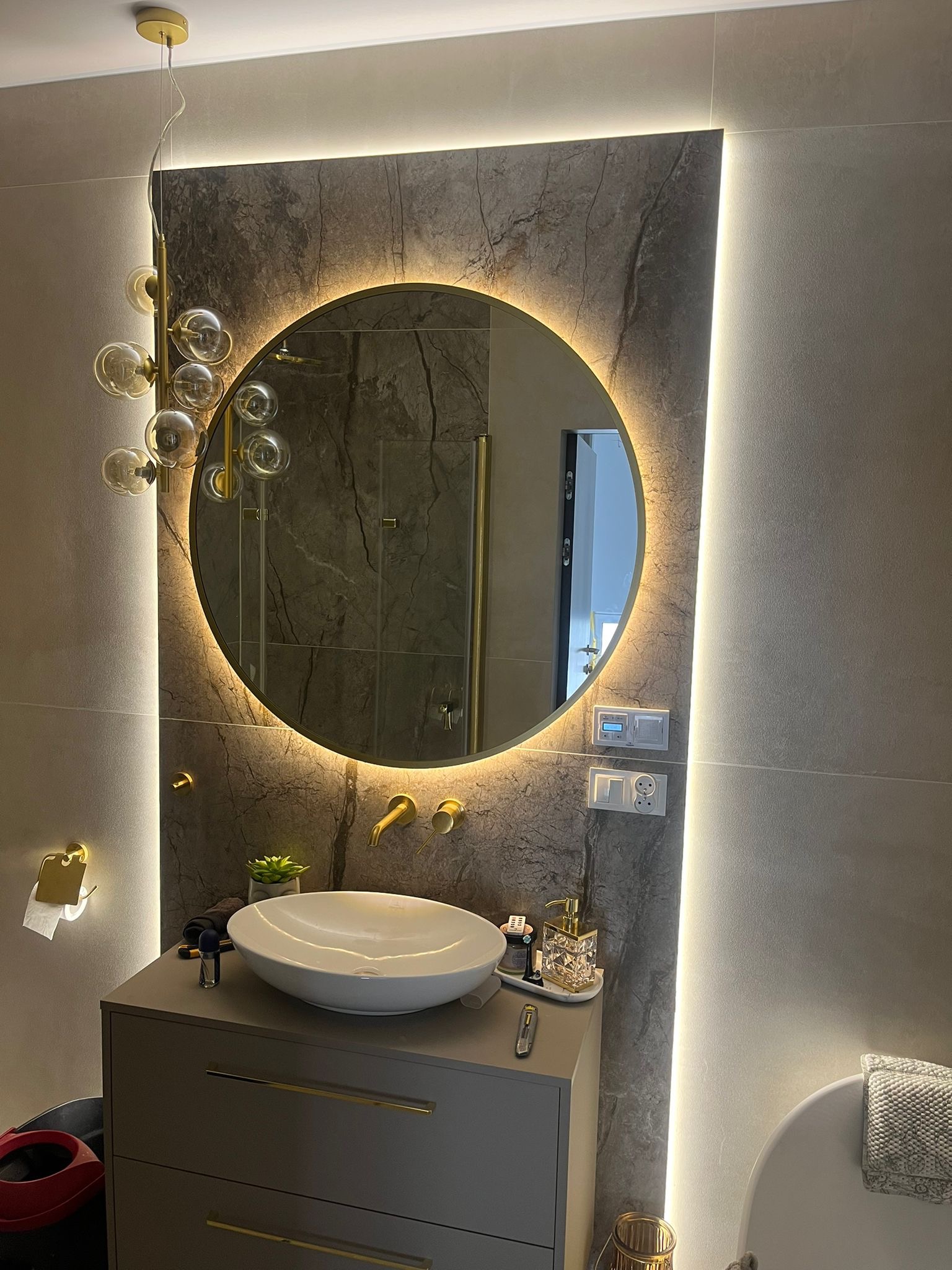 Nowoczesna łazienka z okrągłym lustrem LED, umywalką nablatową, złotą armaturą i dekoracyjnym oświetleniem wiszącym z kulistych kloszy. Ściany wykończone płytami imitującymi marmur z podświetleniem.