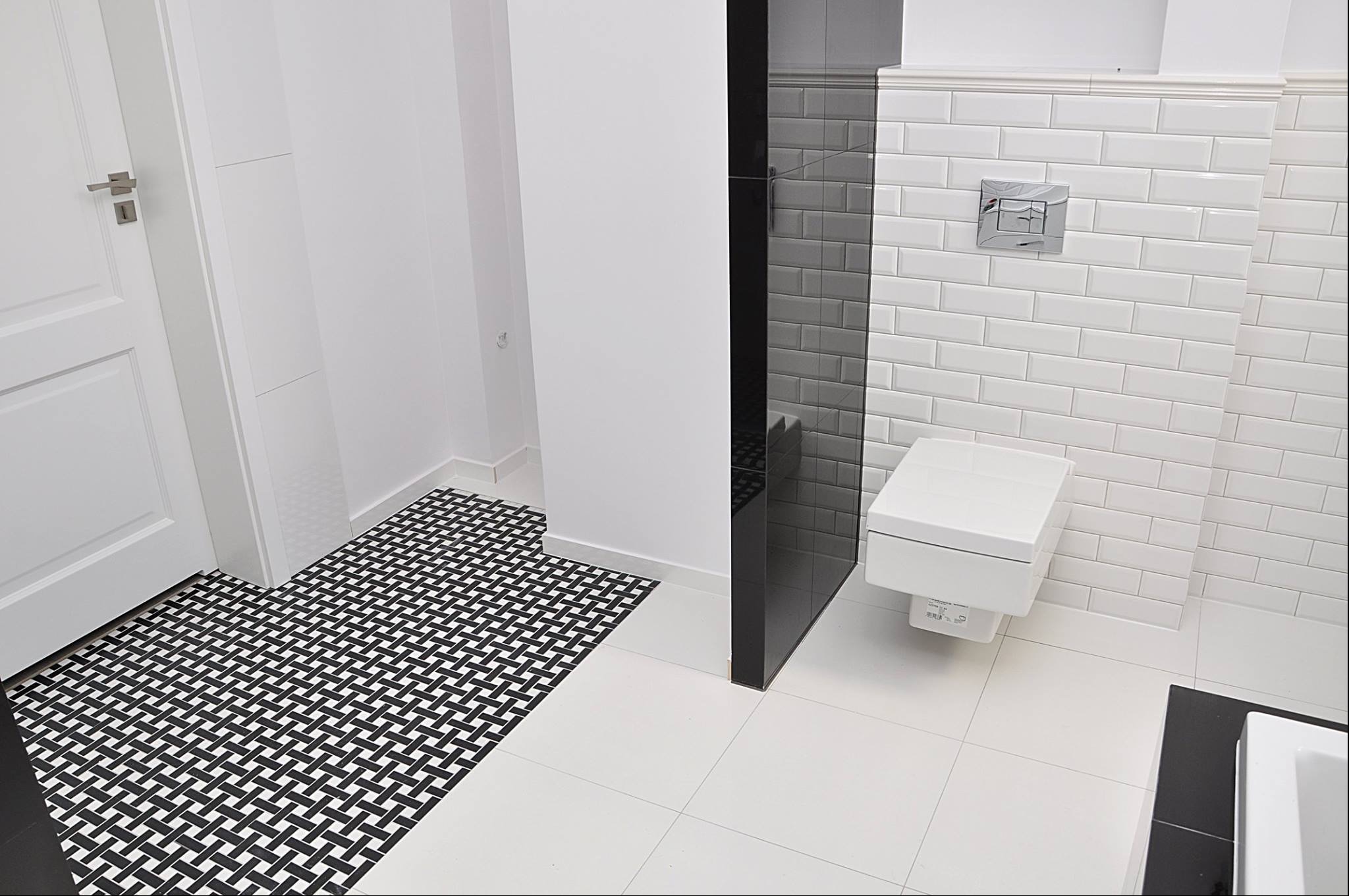 Nowoczesna łazienka z białą wiszącą toaletą, białą cegłą na ścianach, czarnym szklanym panelem i wzorzystą czarno-białą podłogą przy wejściu.