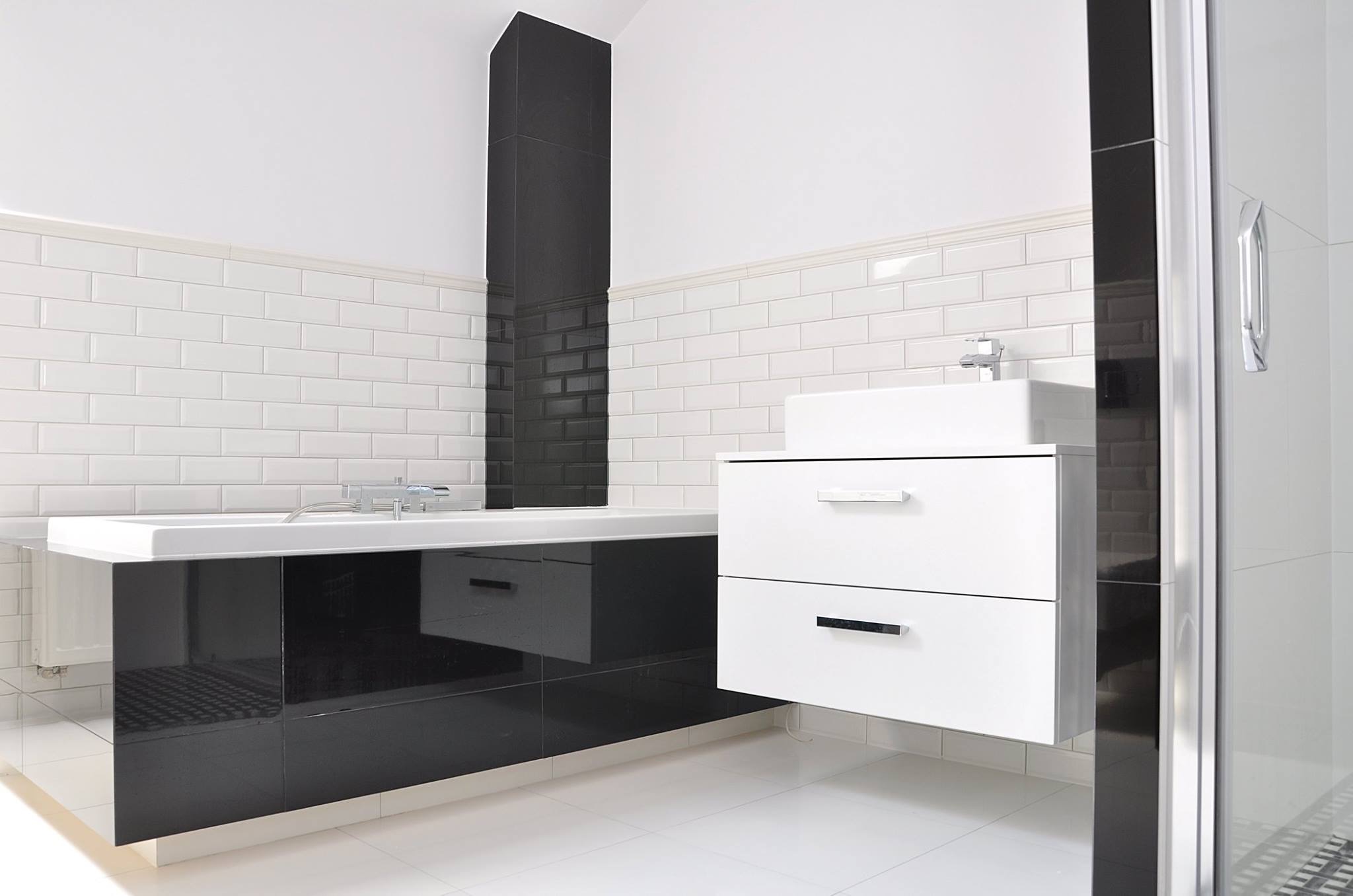 Nowoczesna łazienka w bieli i czerni, z wanną obudowaną czarnym panelem na wysoki połysk, białą umywalką z szafką z dwoma szufladami i biało-czarną ścianą z płytek typu metro.