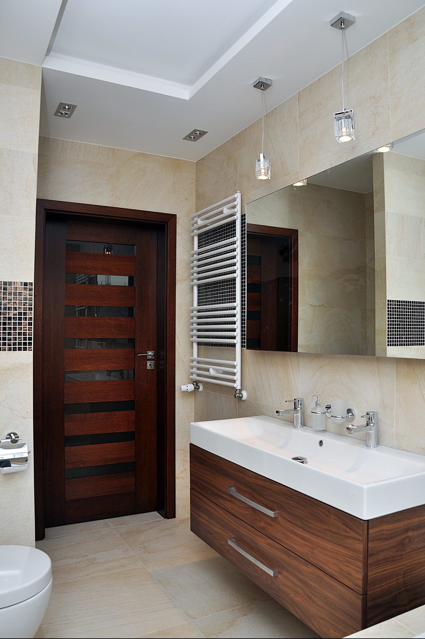 Nowoczesna łazienka z beżowymi płytkami, wiszącą szafką z umywalką z drewnianym frontem, grzejnikiem drabinkowym i ciemnymi drzwiami z przeszkleniami.