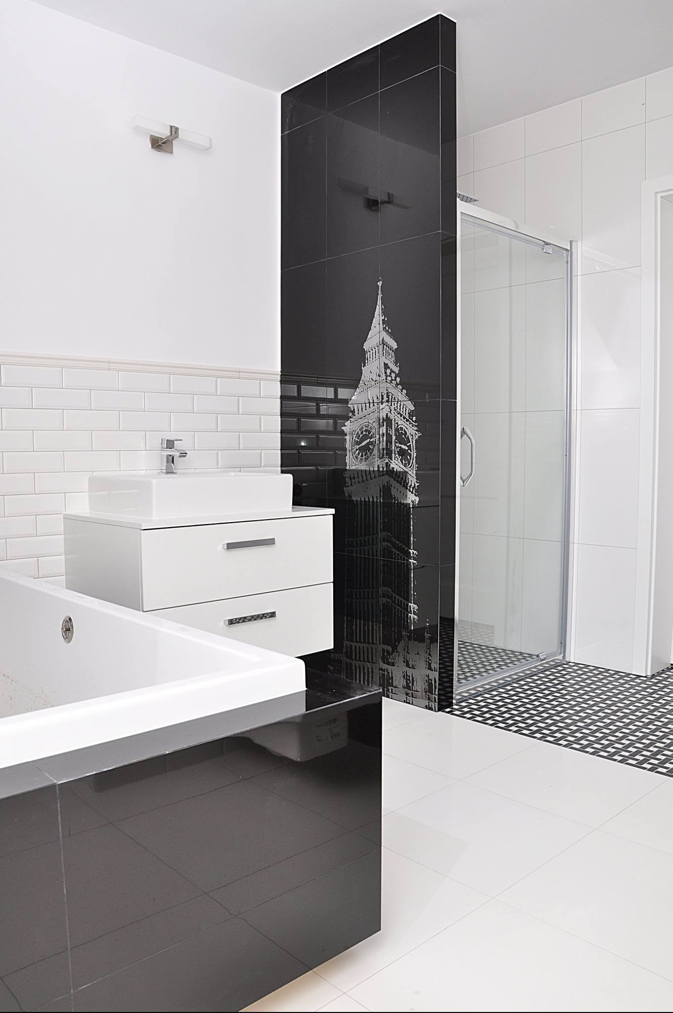Nowoczesna łazienka w bieli i czerni z akcentem londyńskim: czarne płytki z grafiką Big Bena, biała umywalka nablatowa na szafce, kabina prysznicowa i wzorzysta podłoga.