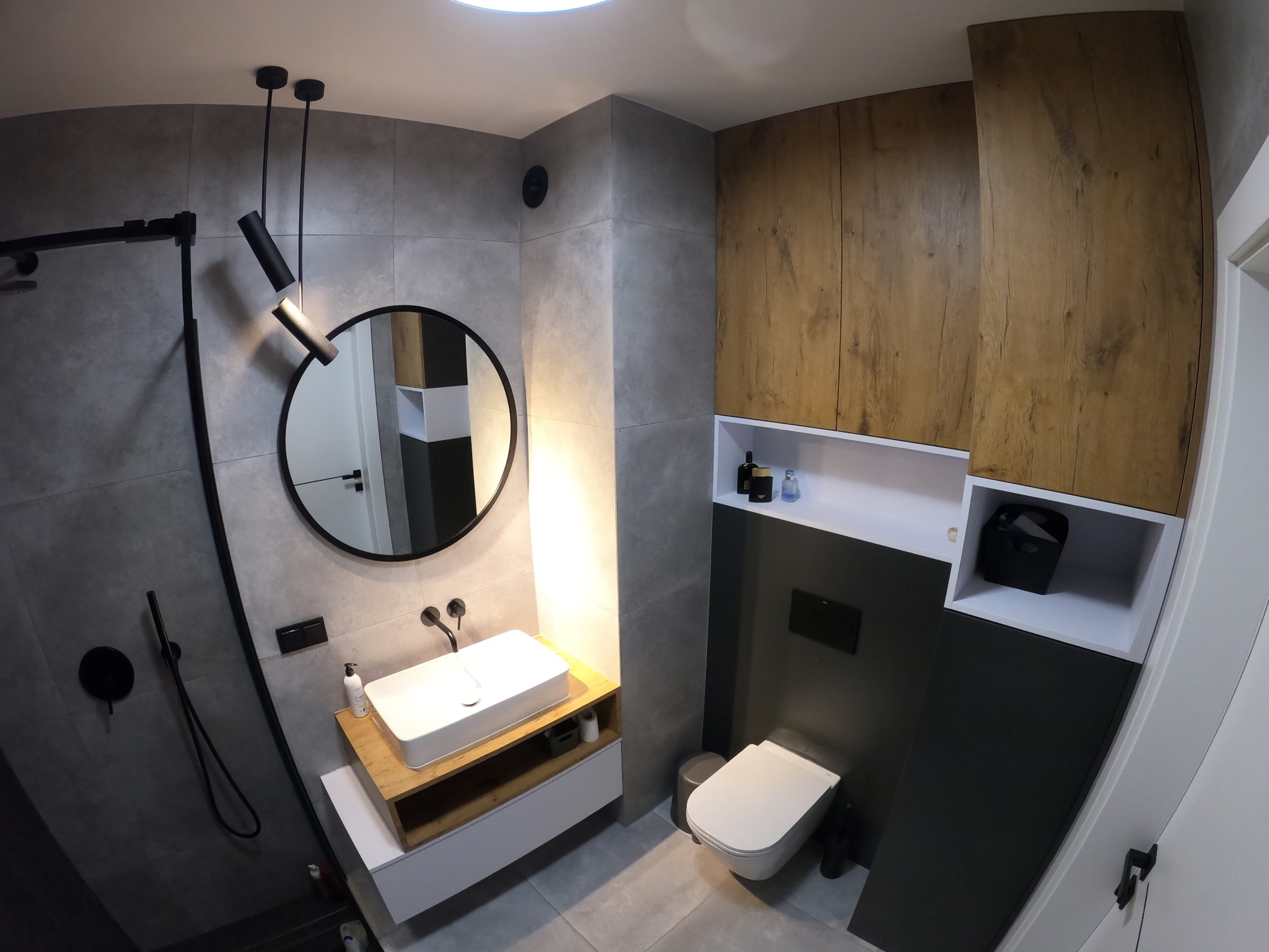 Nowoczesna łazienka z szarymi płytkami, drewnianymi szafkami i okrągłym lustrem, oświetlona wiszącymi lampami punktowymi
