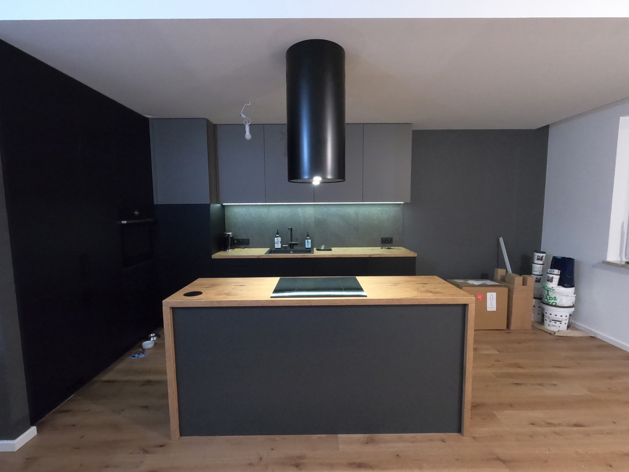 Nowoczesna kuchnia z wyspą, czarne i szare szafki, drewniany blat, minimalistyczny okap cylindryczny, widoczne puszki farb w tle.
