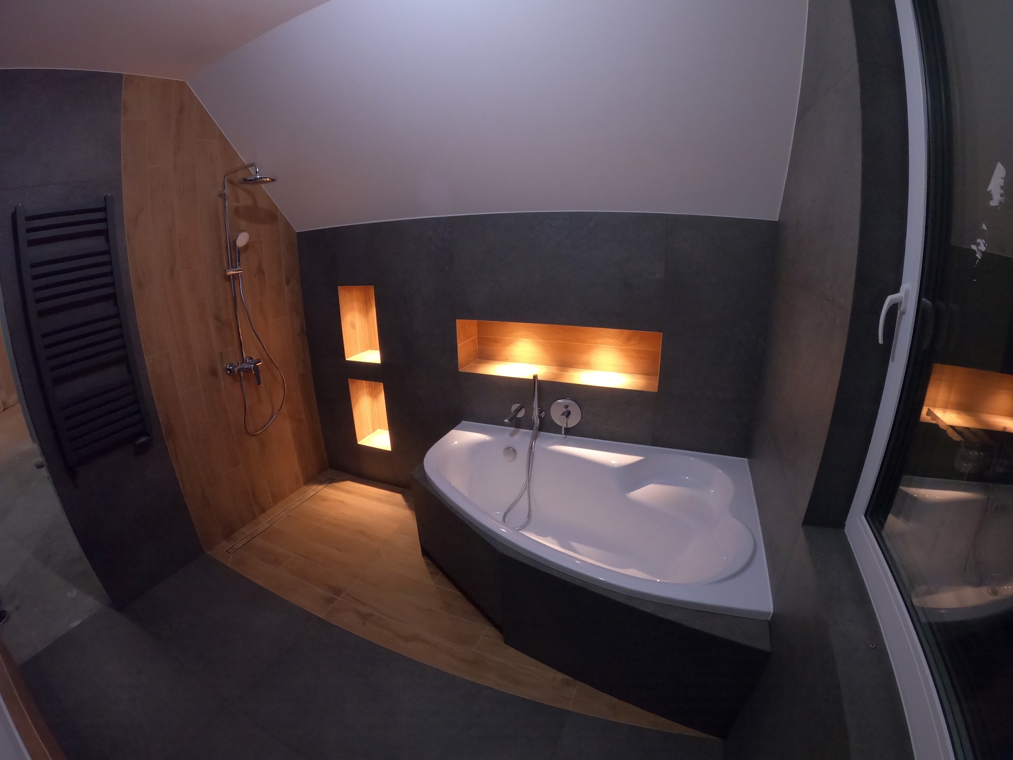 Nowoczesna łazienka z narożną wanną, drewnopodobnymi płytkami i wnękami oświetlonymi ciepłym światłem, widok z szerokiego kąta.