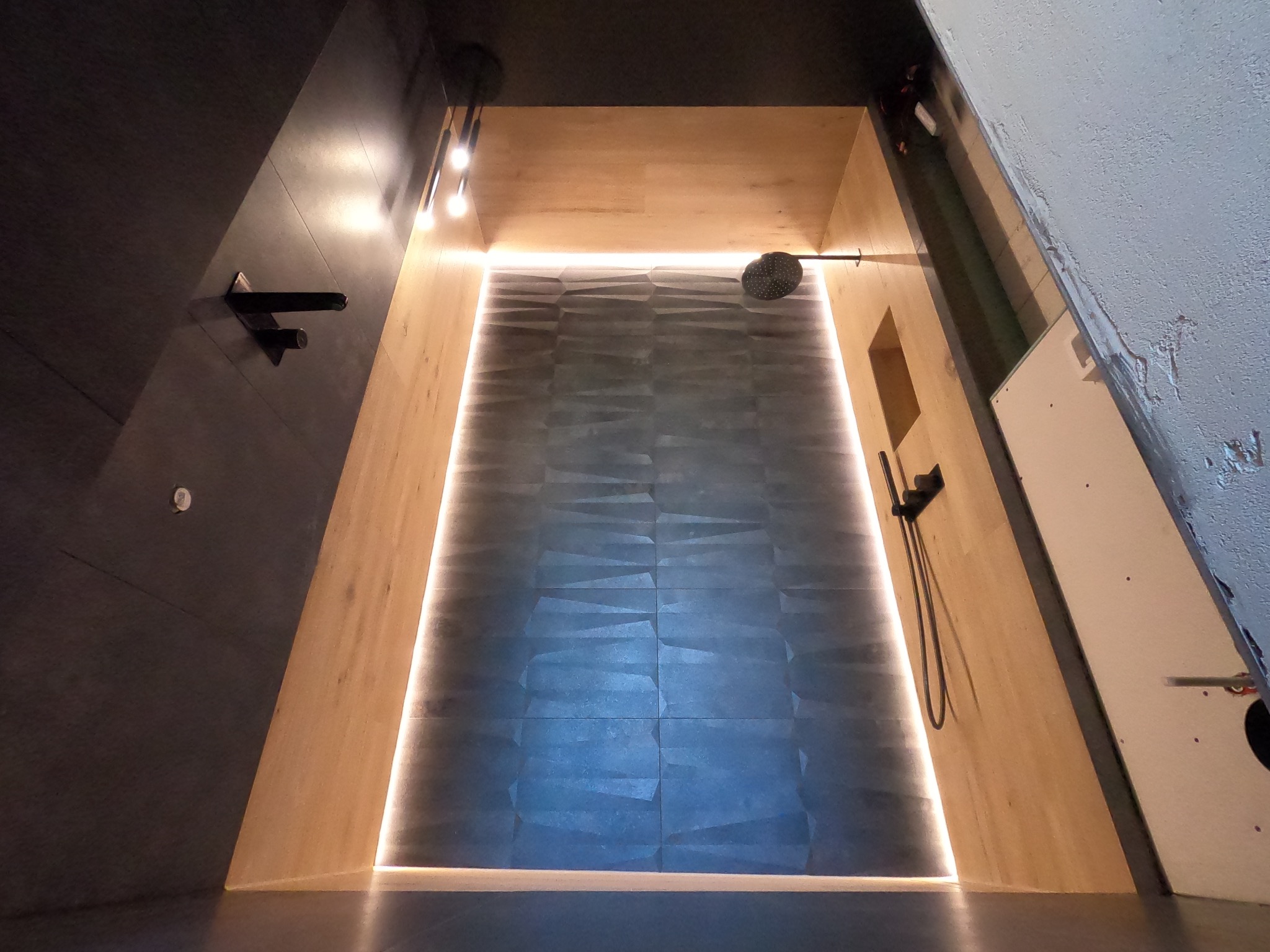 Nowoczesna kabina prysznicowa z podświetleniem LED, wykończona drewnem i ciemnymi płytkami z geometrycznym wzorem, widok od dołu.