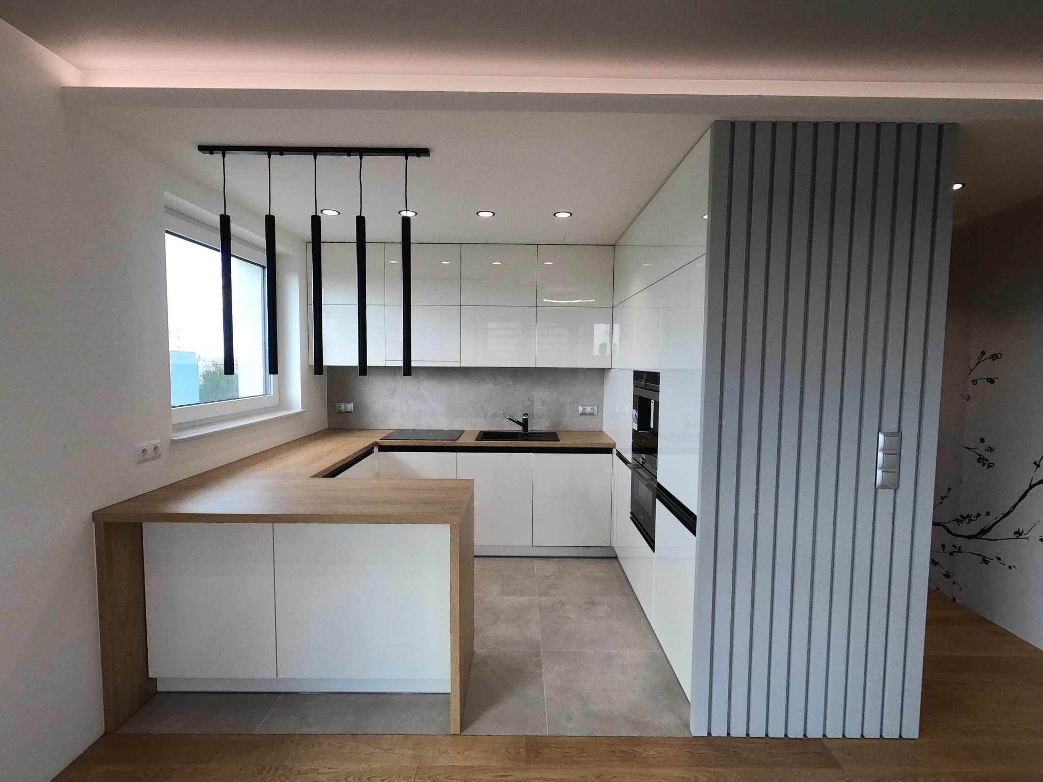 Nowoczesna, jasna kuchnia z białymi szafkami na wysoki połysk, drewnianym blatem i podłogą, oświetlona wiszącymi lampami tubowymi i sufitowymi punktami świetlnymi, z dekoracyjnym panelem ściennym.