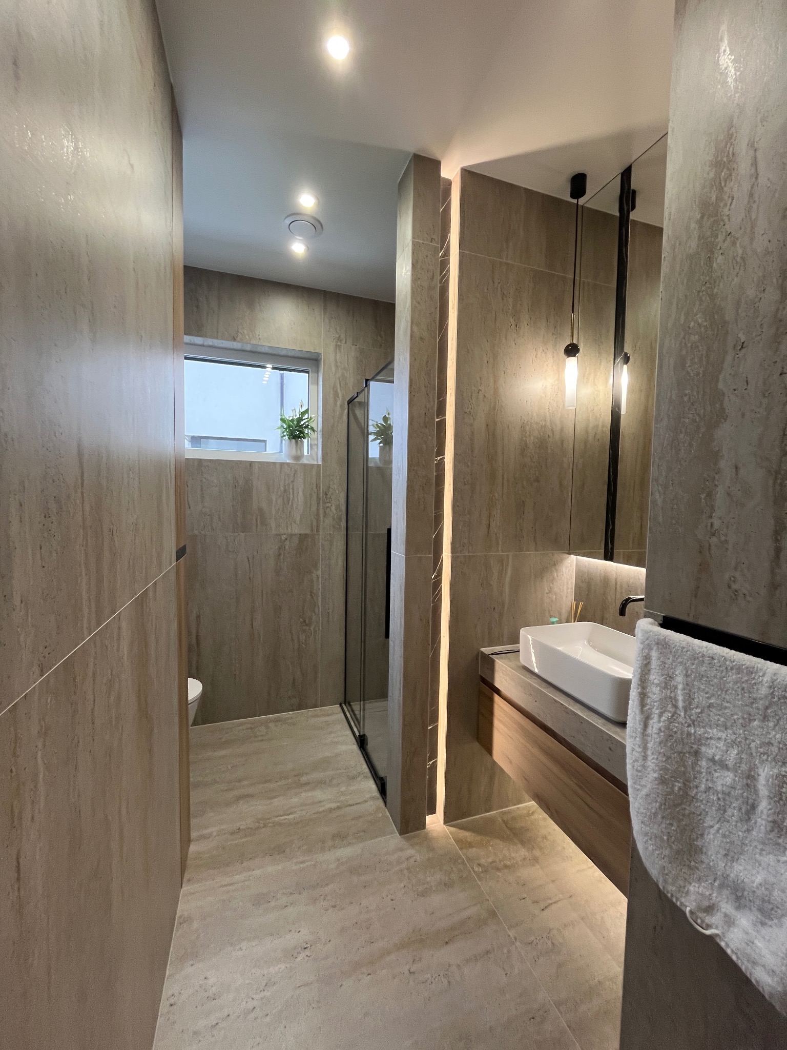 Nowoczesna łazienka w odcieniach beżu z kabiną prysznicową typu walk-in, prostokątną umywalką nablatową, podświetlanym lustrem i wiszącą lampą. Na parapecie okna doniczka z zieloną rośliną.