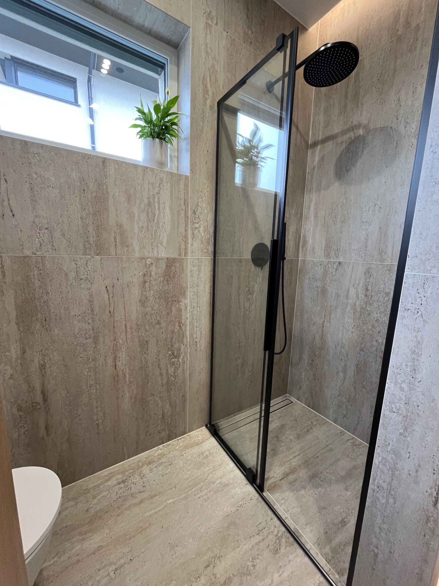 Nowoczesna łazienka z kabiną prysznicową typu walk-in z czarnymi profilami, beżowe płytki imitujące trawertyn na ścianach i podłodze, roślina doniczkowa na parapecie okna.
