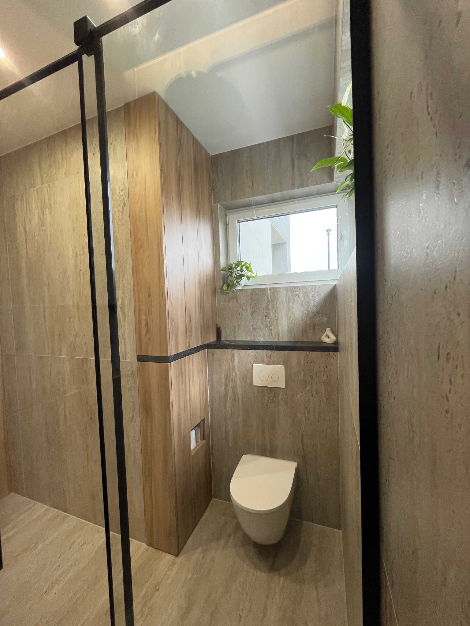 Nowoczesna łazienka z kabiną prysznicową z czarnymi ramami, podwieszana toaleta, płytki imitujące drewno na ścianie, mała półka pod oknem z dekoracją i roślina w doniczce.