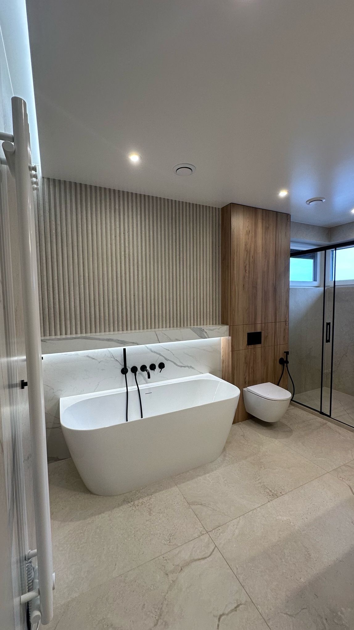 Nowoczesna łazienka z wolnostojącą wanną, czarną armaturą, podświetlanym marmurowym panelem i kabiną prysznicową ze szklanymi drzwiami.