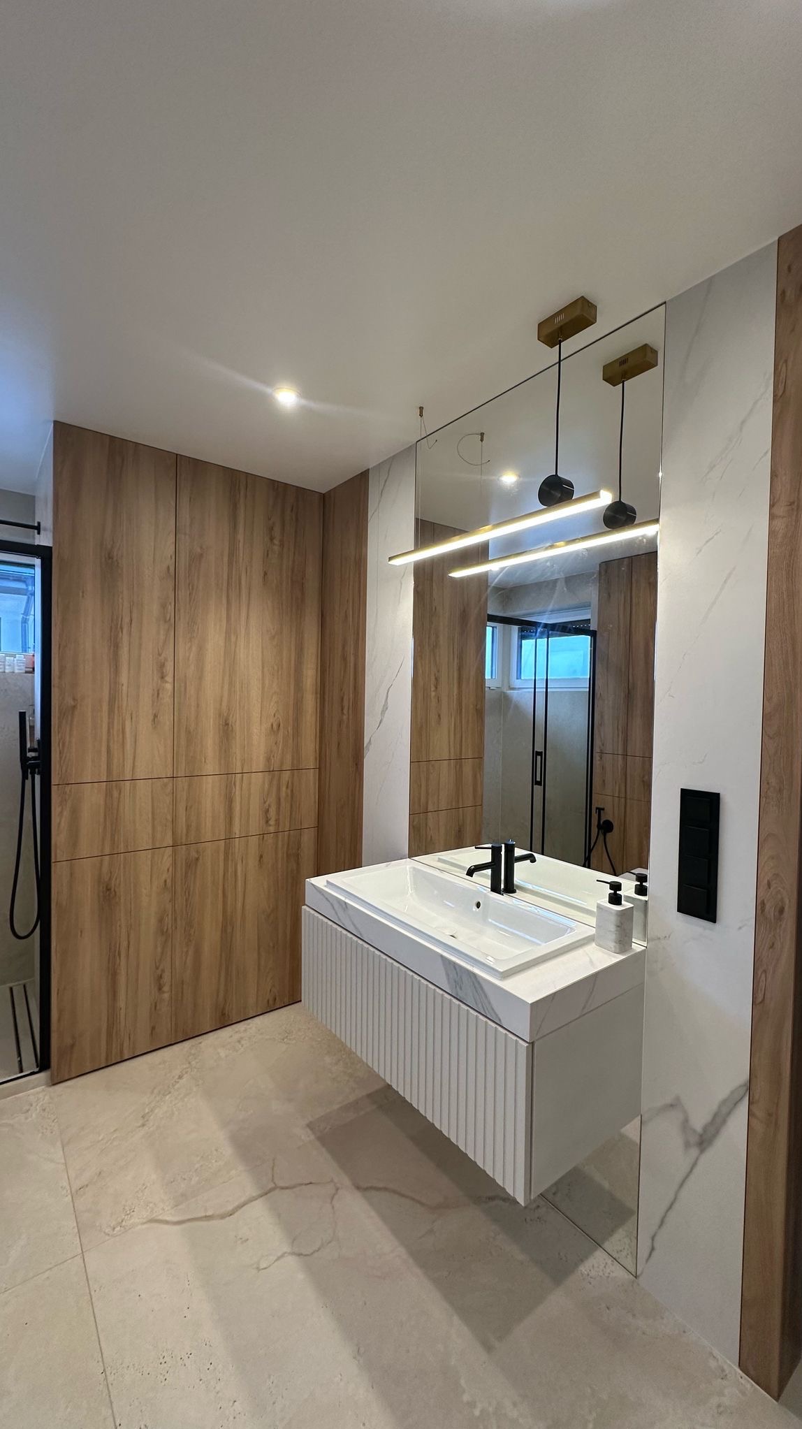 Nowoczesna łazienka z drewnianą zabudową, umywalką nablatową z szafką z żłobieniami, lustrem z podświetleniem LED i czarną armaturą.