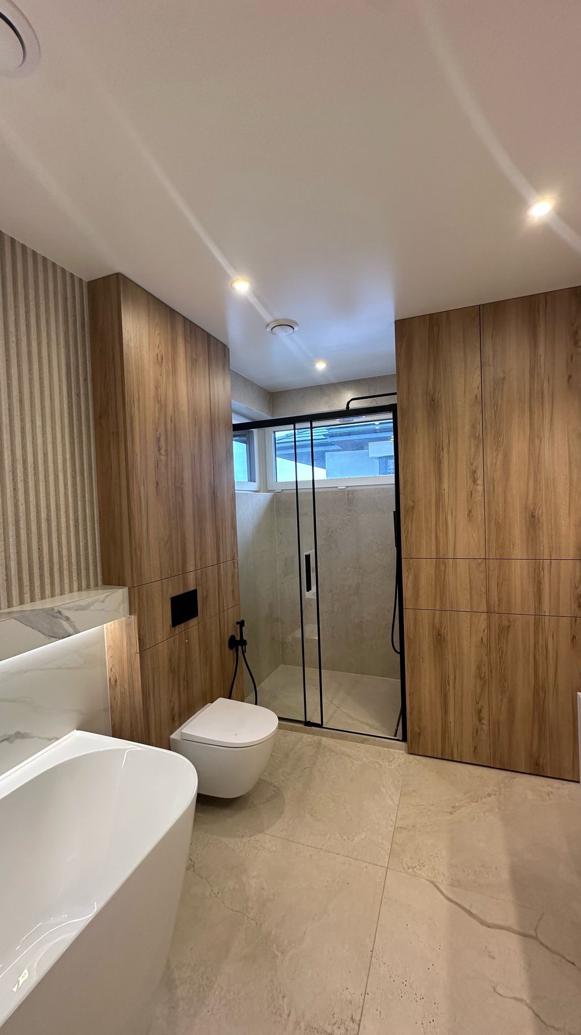 Nowoczesna łazienka z kabiną prysznicową z czarnymi ramami, wiszącą toaletą, wolnostojącą wanną i okładziną ścienną imitującą drewno.