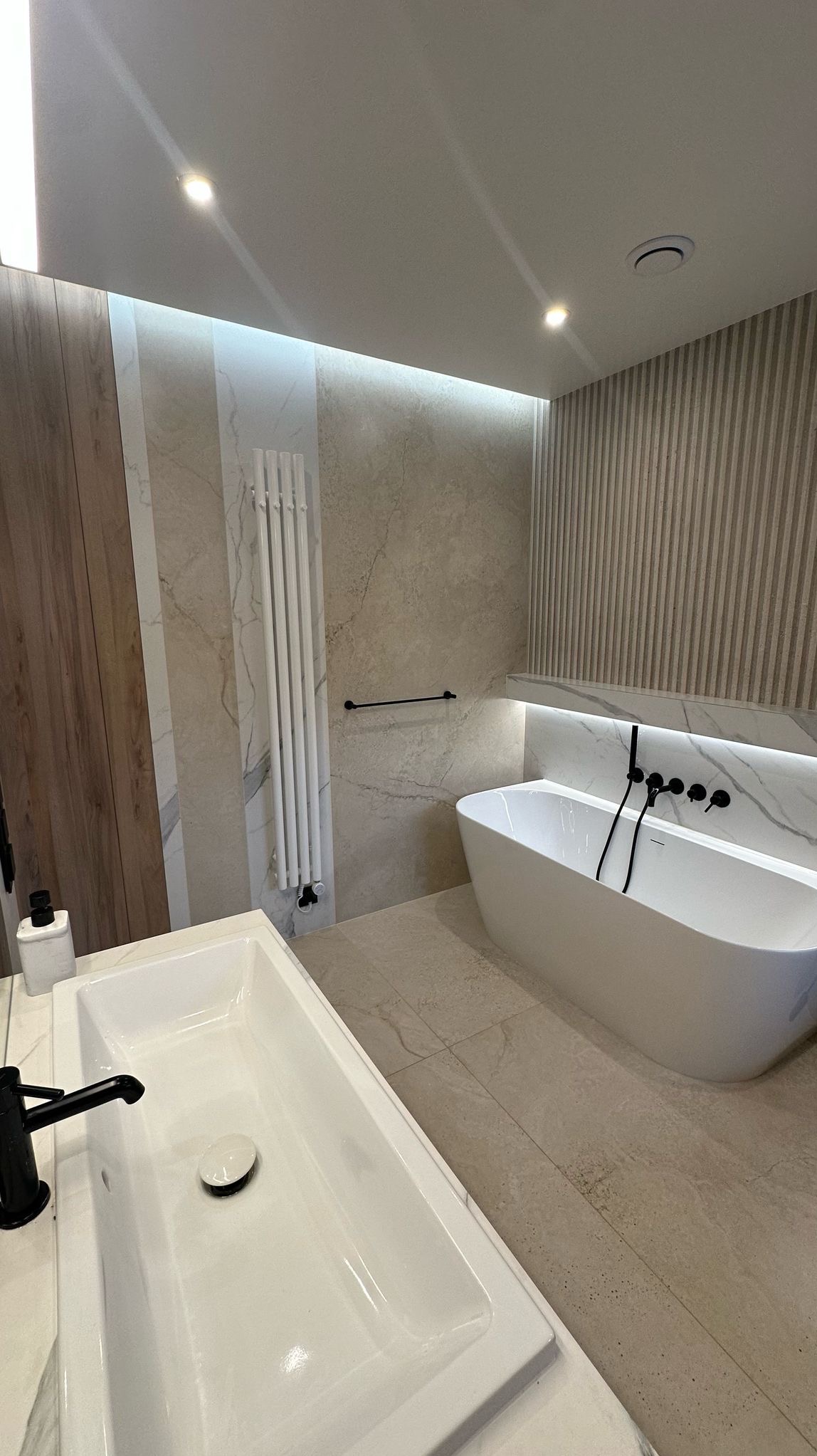 Nowoczesna łazienka z wolnostojącą wanną, białą umywalką nablatową z czarną armaturą i pionowym grzejnikiem drabinkowym na tle beżowych płytek imitujących marmur.