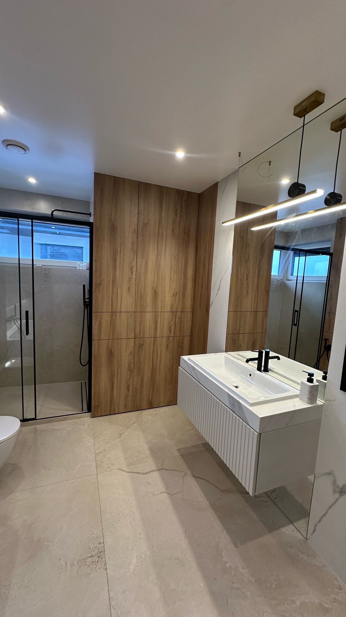 Nowoczesna łazienka z kabiną prysznicową typu walk-in z czarnymi ramami, drewnianą zabudową meblową, podwieszaną szafką z umywalką z blatem imitującym marmur i minimalistycznym oświetleniem.