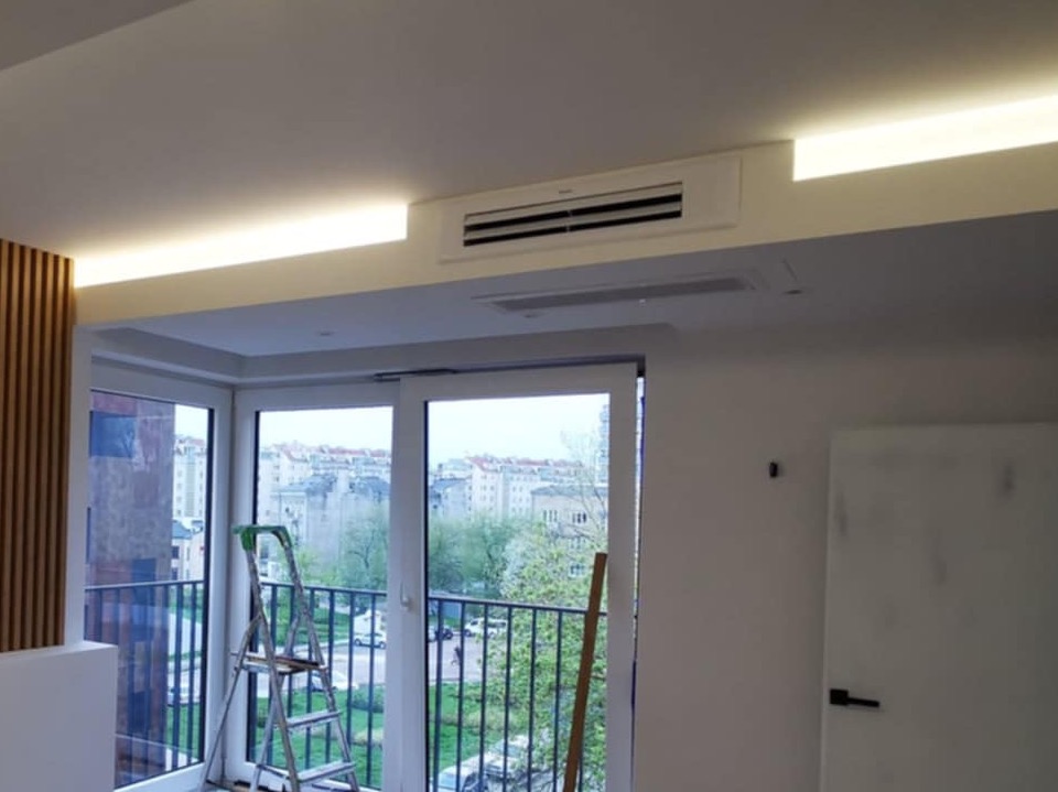 Wnętrze mieszkania w trakcie montażu klimatyzacji kanałowej, widoczna drabina oparta o balkon, obok drewniane panele dekoracyjne i listwy LED w suficie.