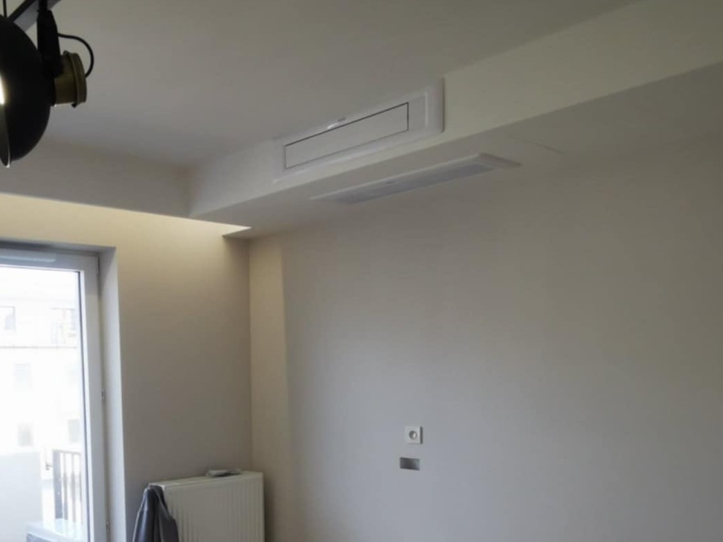 Nowoczesny system klimatyzacji kanałowej w suficie podwieszanym, widoczny radiator, gładkie ściany, szara zasłona na grzejniku, lampa przemysłowa na suficie.