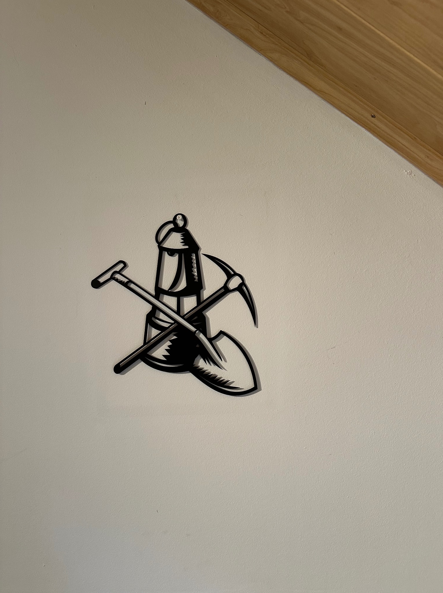 Metalowa dekoracja ścienna przedstawiająca skrzyżowane narzędzia górnicze: kilof, łopatę i lampę górniczą, umieszczona na gładkiej, białej ścianie pod drewnianym sufitem.