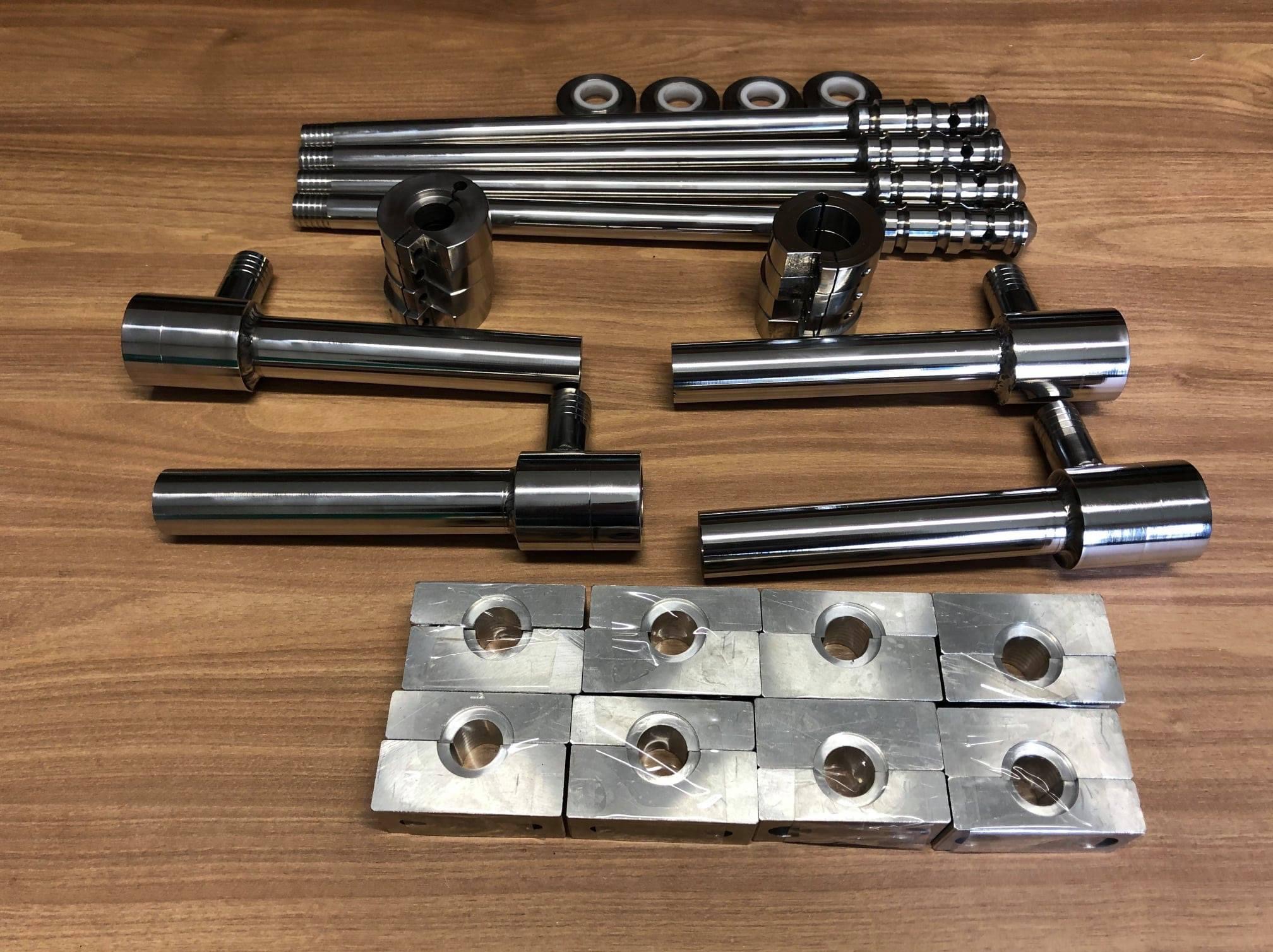 Zestaw precyzyjnie obrobionych, chromowanych elementów metalowych, w tym rur, tulei, i łączników z gwintowanymi końcówkami, ułożonych na drewnianej powierzchni.