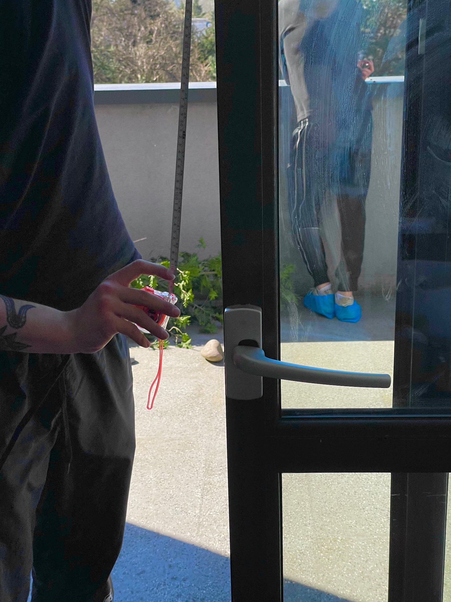 Pomiar okna przed montażem, widoczna miarka w dłoni, na zewnątrz osoba w ochraniaczach na buty.