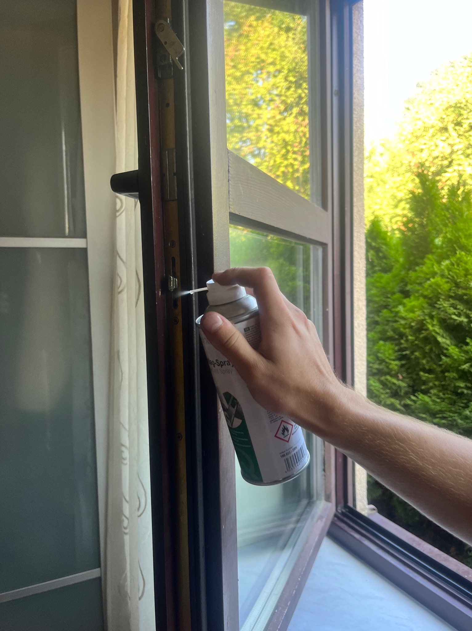 Konserwacja okna: dłoń aplikuje smar w sprayu na mechanizm okienny przy otwartym oknie, z widocznym fragmentem firanki i zielenią za oknem.