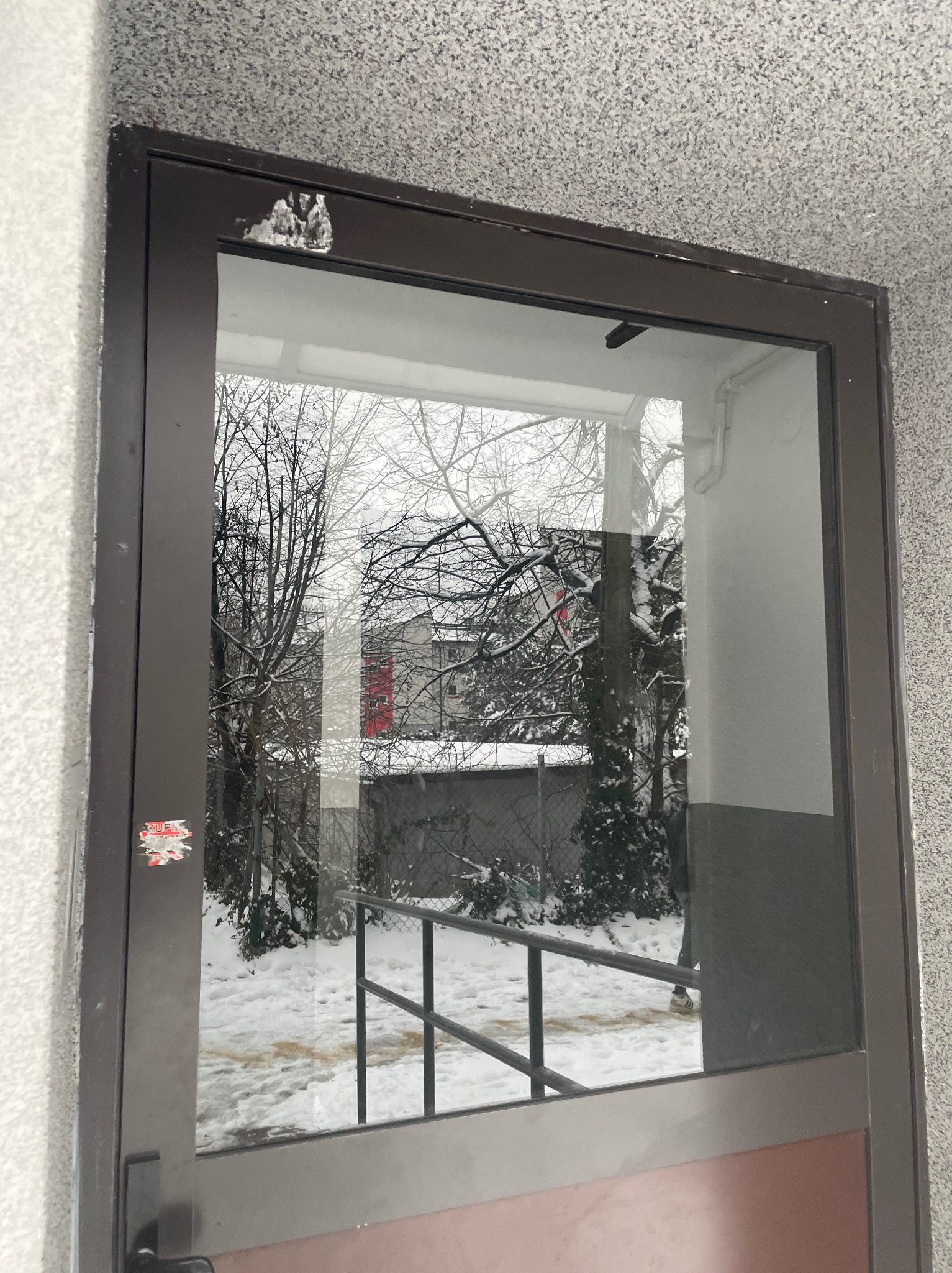 Brązowa rama okienna z naklejką w lewym dolnym rogu, przez szybę widoczny ośnieżony krajobraz z drzewami i budynkiem z czerwoną ścianą w tle.