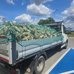 Dominik Wojtaszewski - Ciężarówka Ducato Max wypełniona drewnem kominkowym, zabezpieczonym zieloną siatką, zaparkowana na parkingu w słoneczny dzień. Widoczne błękitne niebo z chmurami.