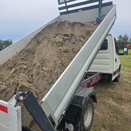 Dominik Wojtaszewski - Ciężarówka z otwartą skrzynią ładunkową, wypełnioną piaskiem, na tle zieleni. Widoczny mechanizm podnoszenia skrzyni i logo firmy na błotniku.
