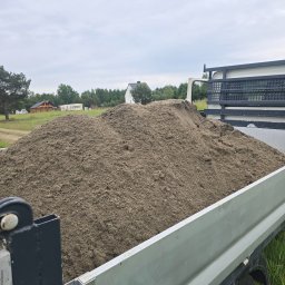 Dominik Wojtaszewski - Przyczepa wypełniona piaskiem na tle zieleni i zabudowań. Widok z boku, ukazujący ładunek i otoczenie. Szary odcień piasku kontrastuje z zielenią.