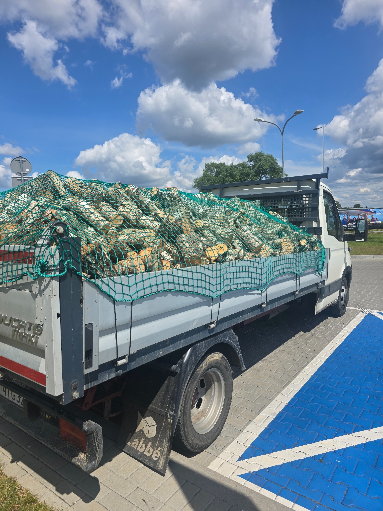 Ciężarówka Ducato Max wypełniona drewnem kominkowym, zabezpieczonym zieloną siatką, zaparkowana na parkingu w słoneczny dzień. Widoczne błękitne niebo z chmurami.