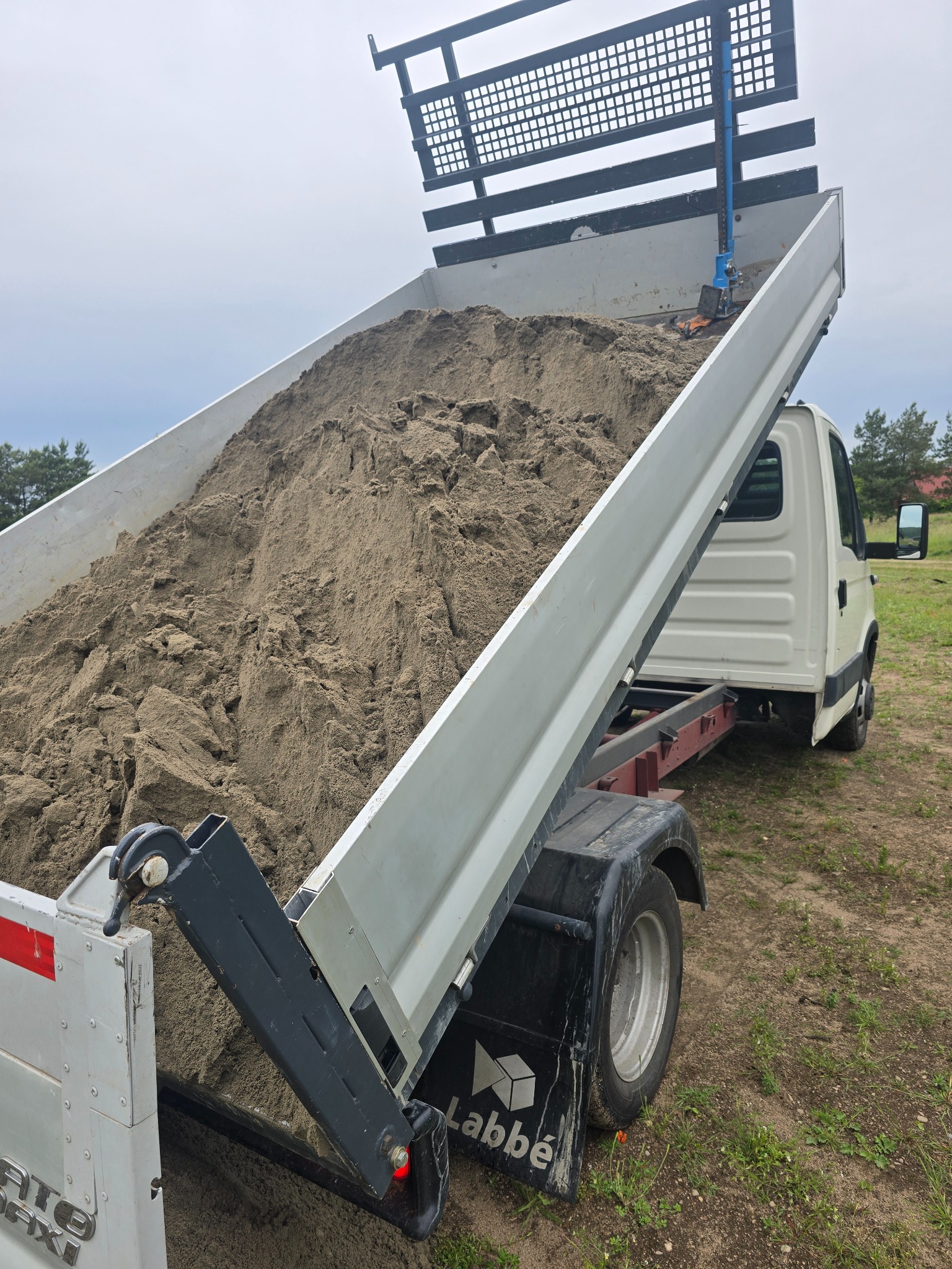 Ciężarówka z otwartą skrzynią ładunkową, wypełnioną piaskiem, na tle zieleni. Widoczny mechanizm podnoszenia skrzyni i logo firmy na błotniku.