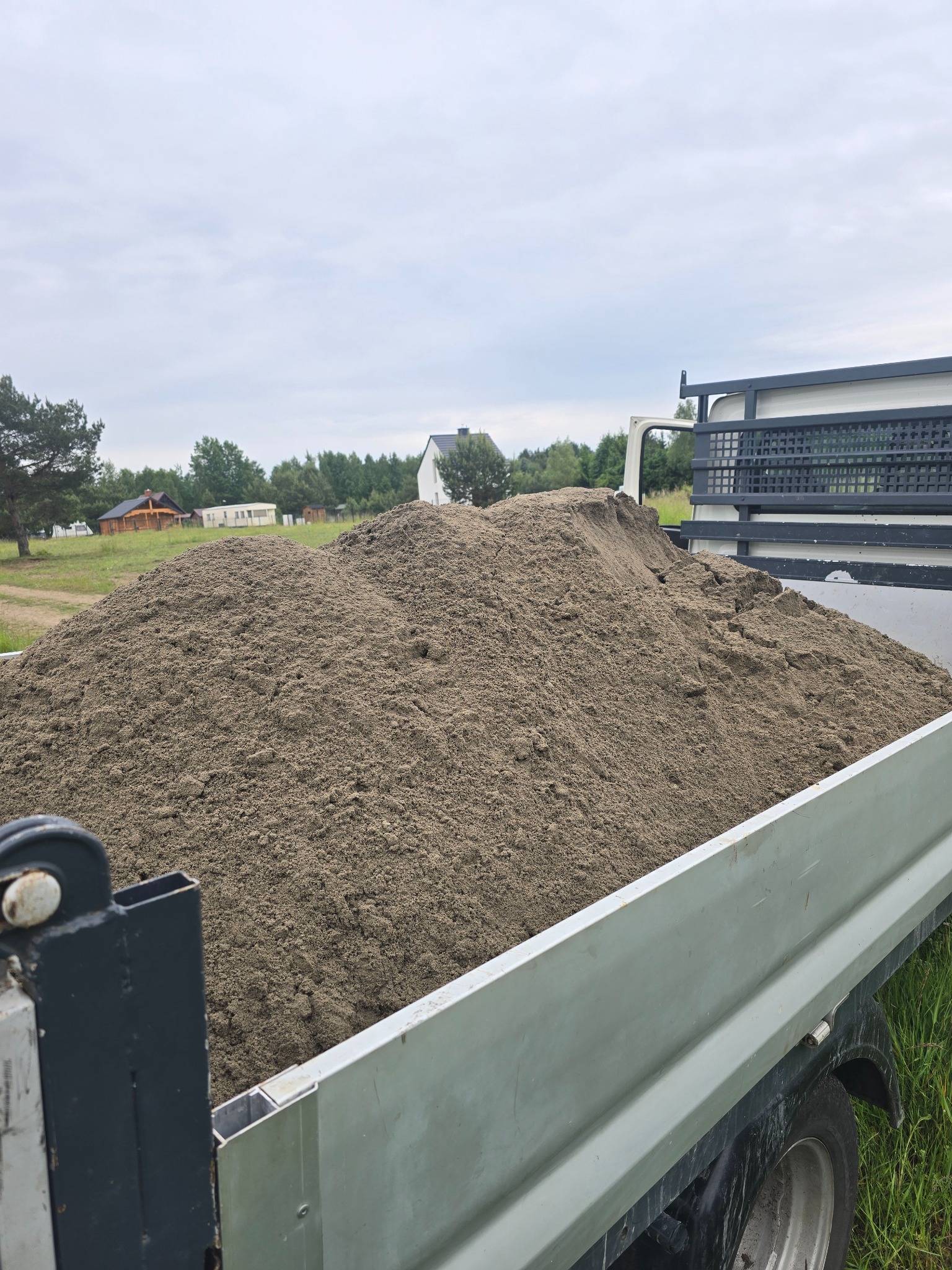 Przyczepa wypełniona piaskiem na tle zieleni i zabudowań. Widok z boku, ukazujący ładunek i otoczenie. Szary odcień piasku kontrastuje z zielenią.