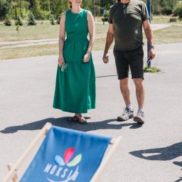 LARSEN - Dwoje ludzi stoi na betonowej powierzchni obok błękitnego leżaka z logo. W tle drzewa i budynki. Jasne światło słoneczne. Kobieta w zielonej sukience.