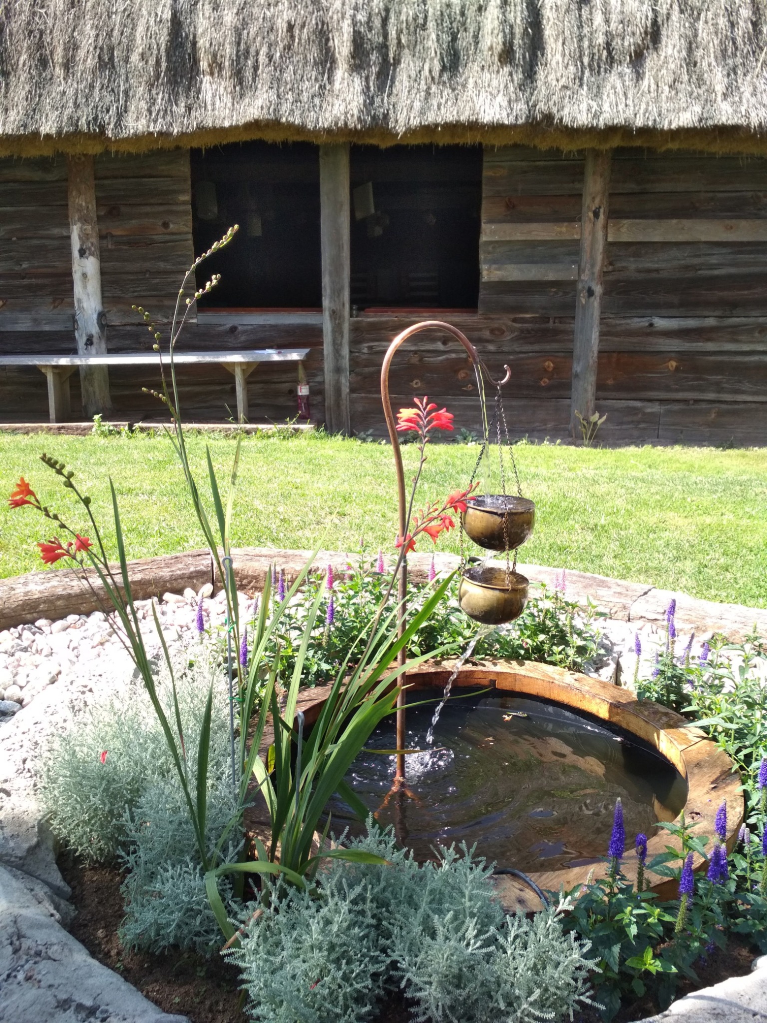 Ogród z małym stawem w drewnianej obudowie i kaskadową fontanną z wiszących donic, na tle drewnianego budynku z trzcinowym dachem.
