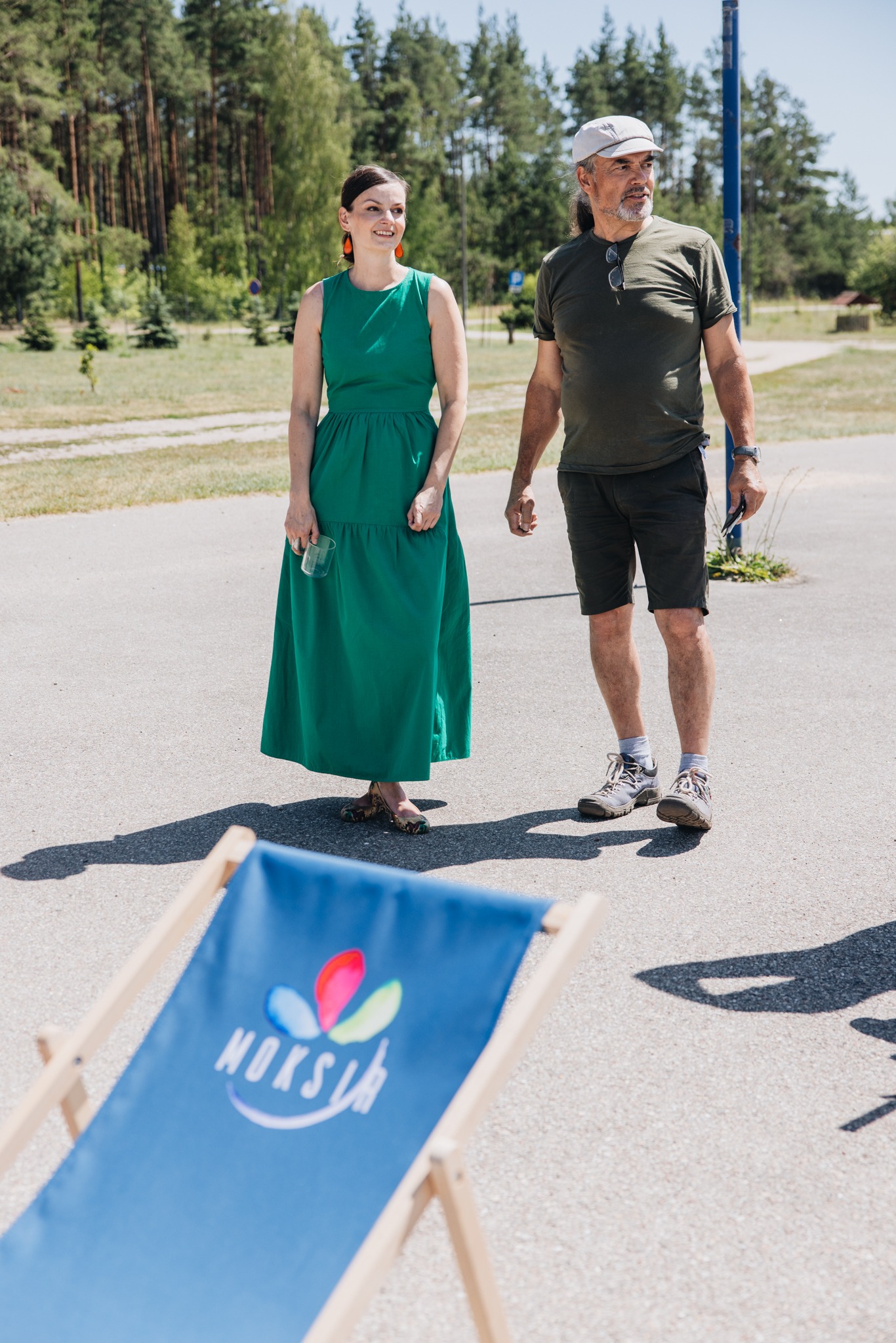 Dwoje ludzi stoi na betonowej powierzchni obok błękitnego leżaka z logo. W tle drzewa i budynki. Jasne światło słoneczne. Kobieta w zielonej sukience.