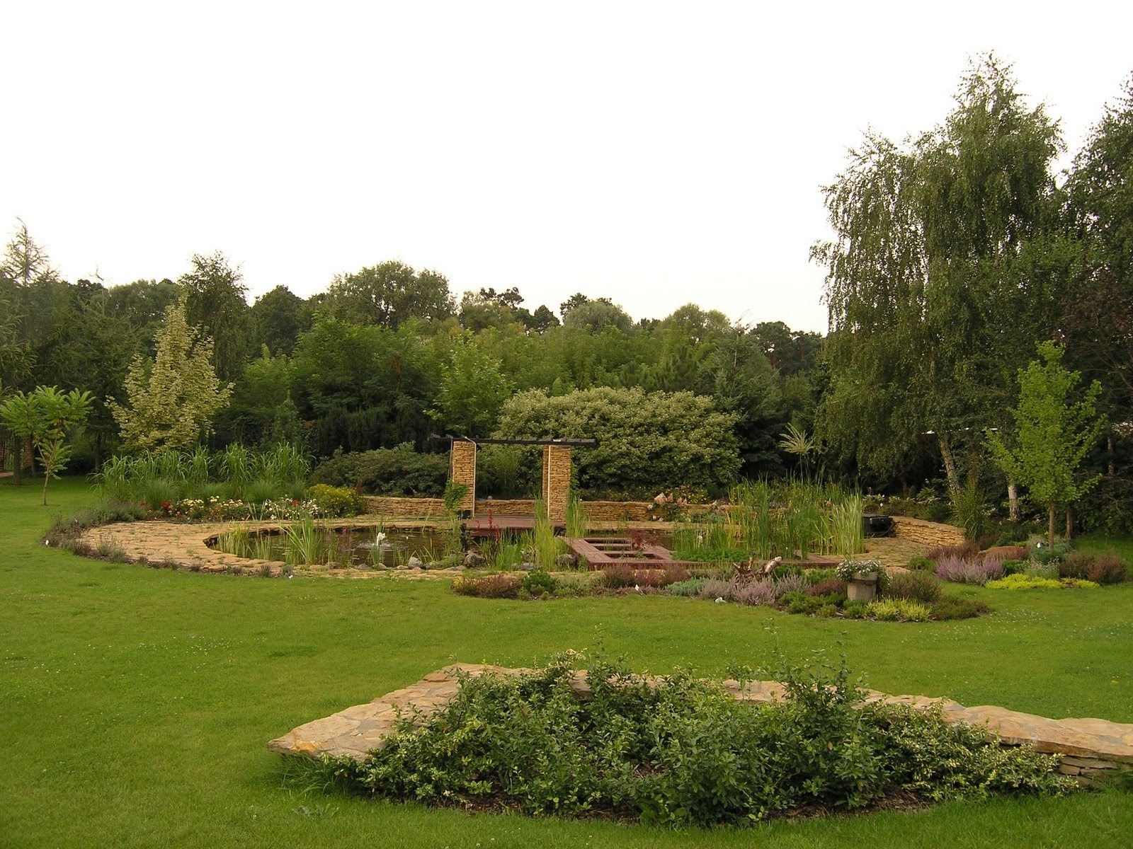 Malowniczy ogród z oczkiem wodnym, kamiennymi ścieżkami i bujną roślinnością. Widoczna drewniana konstrukcja nad wodą i różnorodne gatunki drzew.