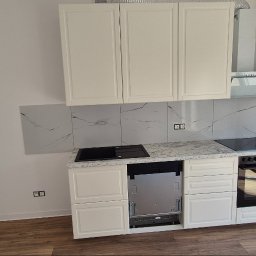 LIBAU Stefan Lizon - Jasna kuchnia z białymi szafkami i marmurowymi płytkami. Widoczna płyta indukcyjna, piekarnik i okap. Nowoczesne wykończenie wnętrza z drewnianą podłogą.