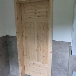 LIBAU Stefan Lizon - Surowe drewniane drzwi z ościeżnicą w nowej łazience z szarymi płytkami podłogowymi i ściennymi, widok na fragment ściany i podłogi, naturalne światło dzienne.