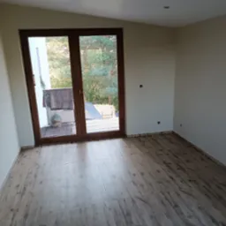 Jasne wnętrze po remoncie z panelami podłogowymi w odcieniach drewna i dużym oknem balkonowym z widokiem na zieleń. Widoczne listwy przypodłogowe.